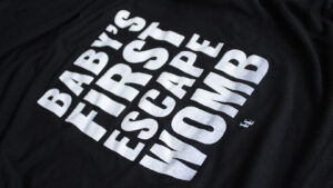 A photo showing a maternity shirt with the phrase Baby's First Escape Womb.